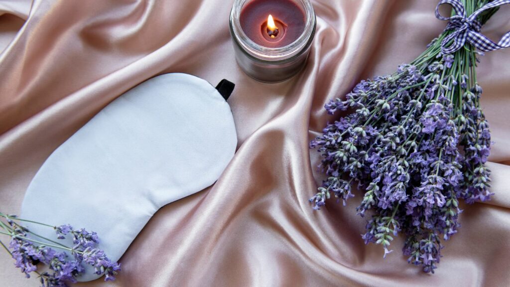 Photo of a sleep mask, some lavender and a candle against a soft pink slik background