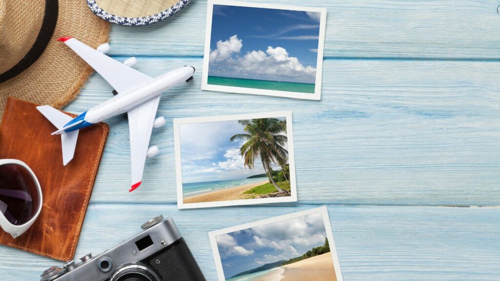 Travel items such as postcards, sunglasses and a camera against a wooden teal backdrop