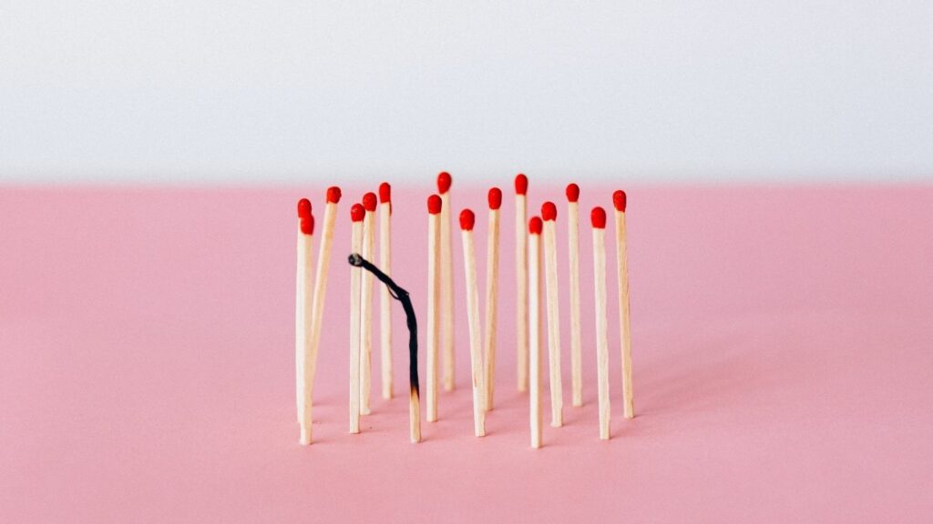 Photo of lots of matches standing on their ends. One of them has been burnt right down to almost the bottom. The matches are against a pale pink background and have red tips.